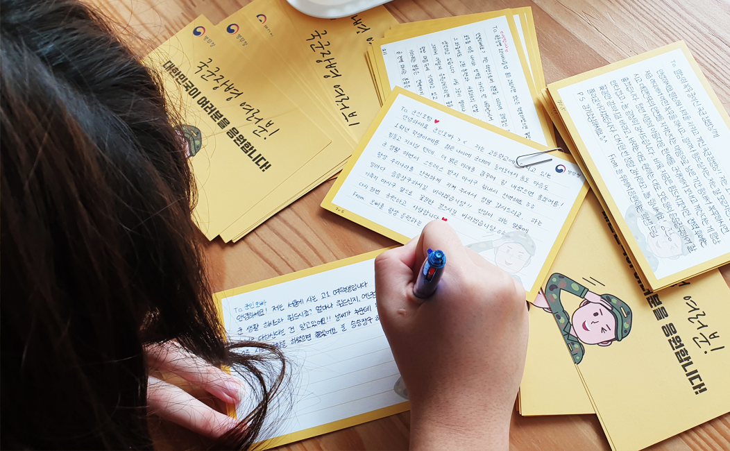편지가 펼쳐져 있는 책상에서 편지를 작성하고 있는 여자아이의 뒷모습