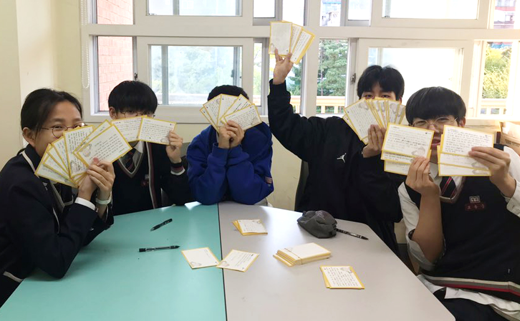 남자, 여자 중학생들이 원탁에 앉아 감사편지로 얼굴을 가리고있는 모습